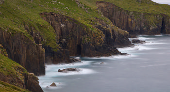 Long exposure Cornish cliffs one This landscape photograph captures the rugged Cornish cliffs in the late afternoon during early summer. The long exposure technique creates a smooth, mist-like effect over the sea as waves wash against the rocky shore, highlighting the dramatic contrast between the solid cliffs and fluid water. The image prominently features the steep cliff faces of Cornwall, covered with patches of grass and dotted with boulders, with a visible cave set into the rock formation. The cave adds depth and character to the scene along the coastline. The overall composition emphasizes the natural beauty and geological features found on the Cornish coast, with the persistent influence of the sea shaping the cliffs and cave over time.